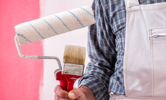 A painter holding a paintbrush and a paint roller in front of a partially painted pink-red wall