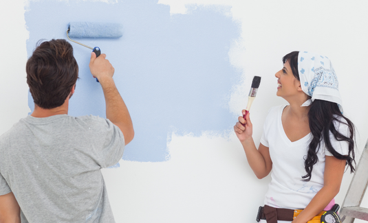 A smiling man and woman laughing together while painting a white interior wall light blue