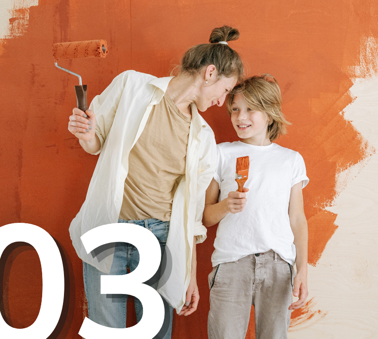 A smiling mother and her young son laughing together while painting an interior wall bright orange